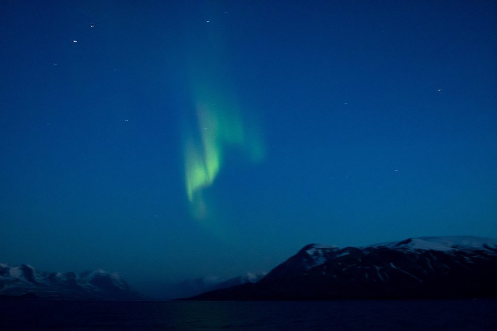 Green northern lights in a blue sky with dark snow covered peaks below. 