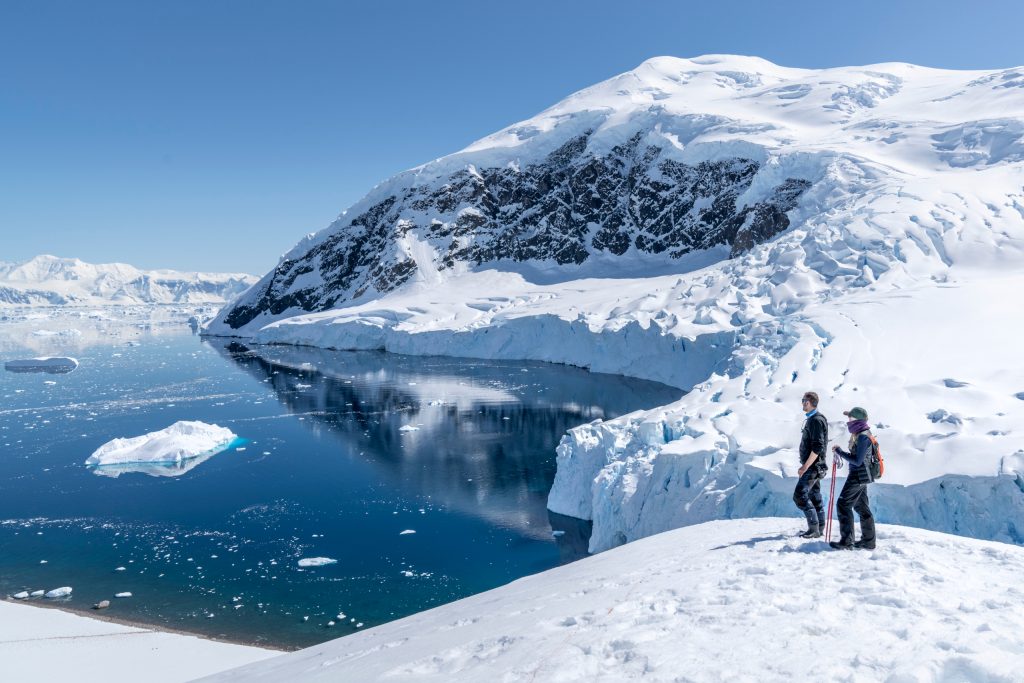 Two guests standing on snowy hillside looking at scenic bay in Antarctica.