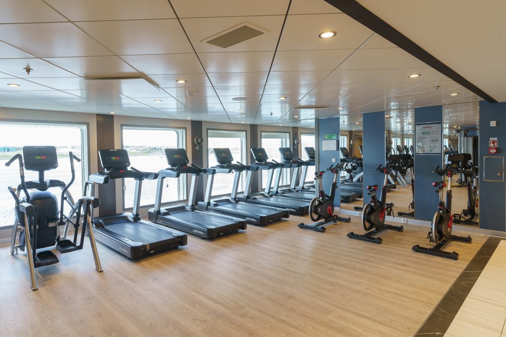 Fitness center with wood floor, four treadmills, elliptical, three bikes and floor to ceiling windows on Ocean Explorer. 