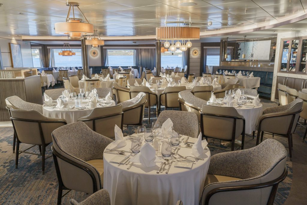 Various circular tables with white tablecloths, chairs, light fixtures and large windows in main dining room on Ocean Explorer. 