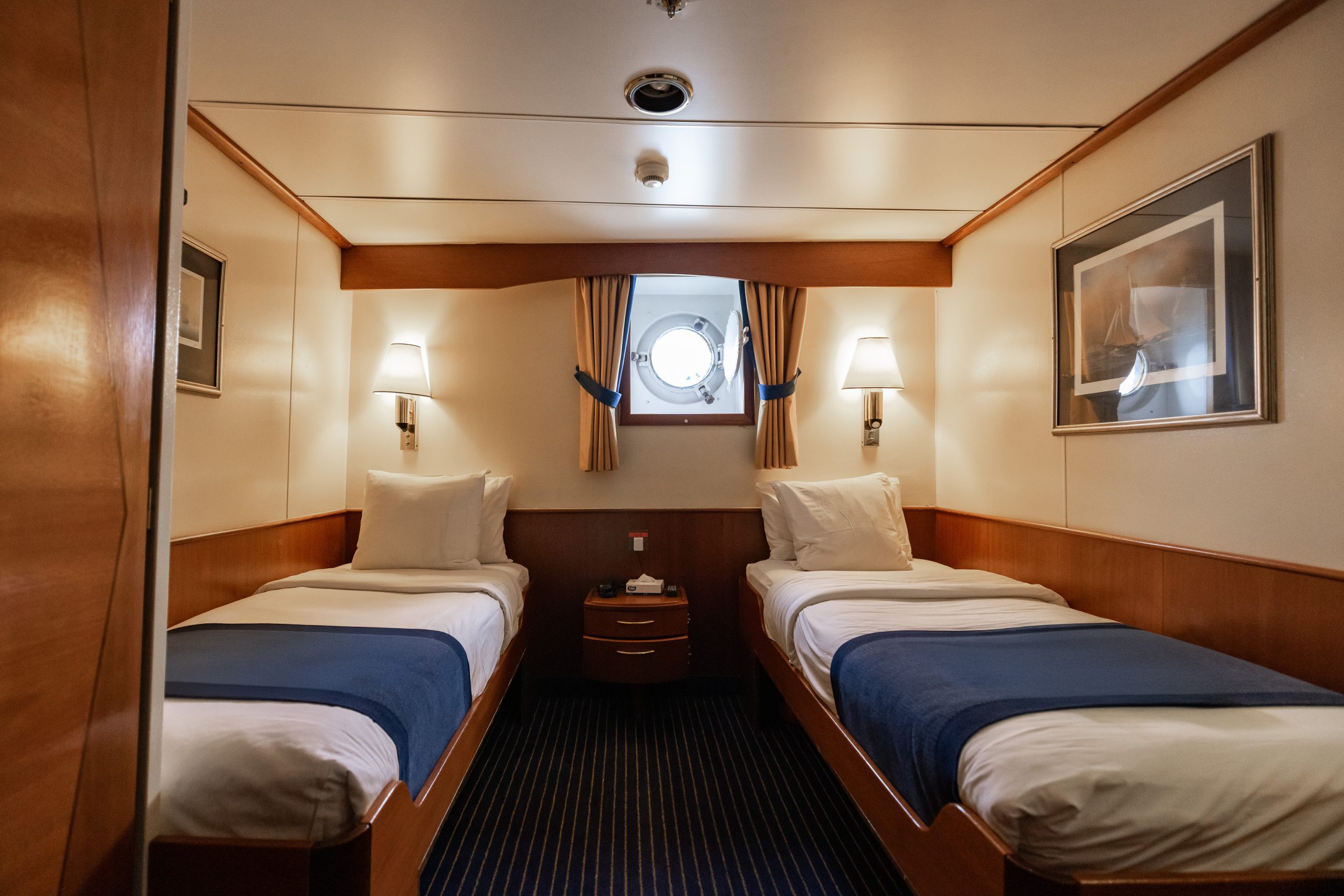 Interior of a Main Deck Twin Porthole cabin on the ship Expedition with 2 twin beds and a small porthole window.
