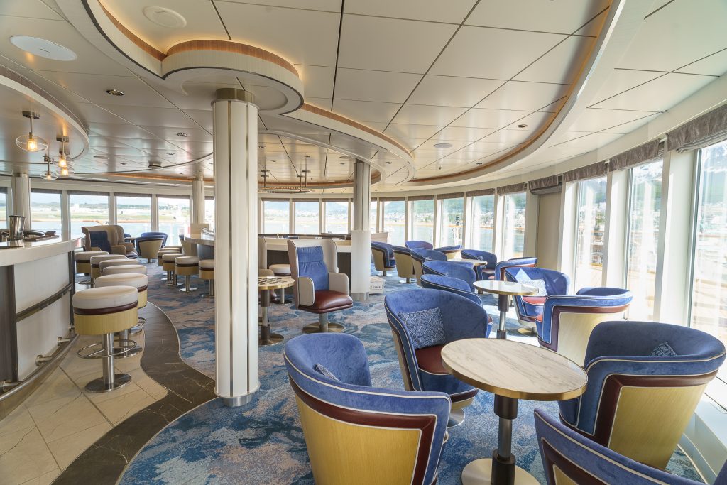 Blue chairs, small tables , white barstools and wrap around windows inside Observation Lounge on Ocean Explorer. 