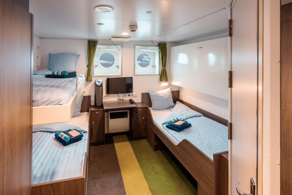 Interior of Triple Porthole cabin with 2 lower berths, 1 upper berth, small desk and 2 porthole windows. 