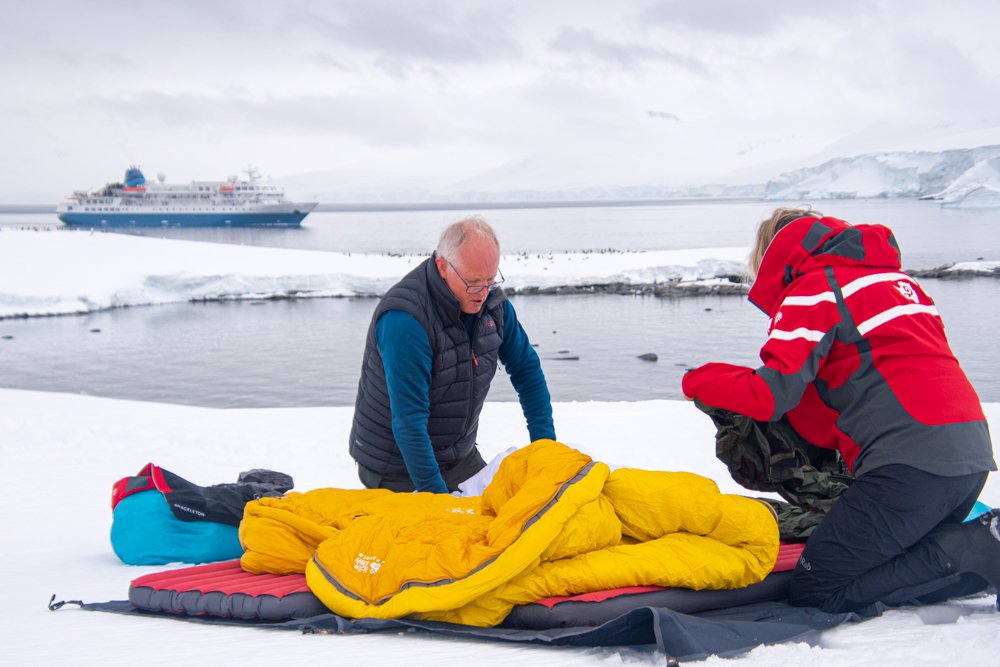 Two campers setting up their sleeping bags on the snow in Antarctica with the Seaventure ship in the background. 