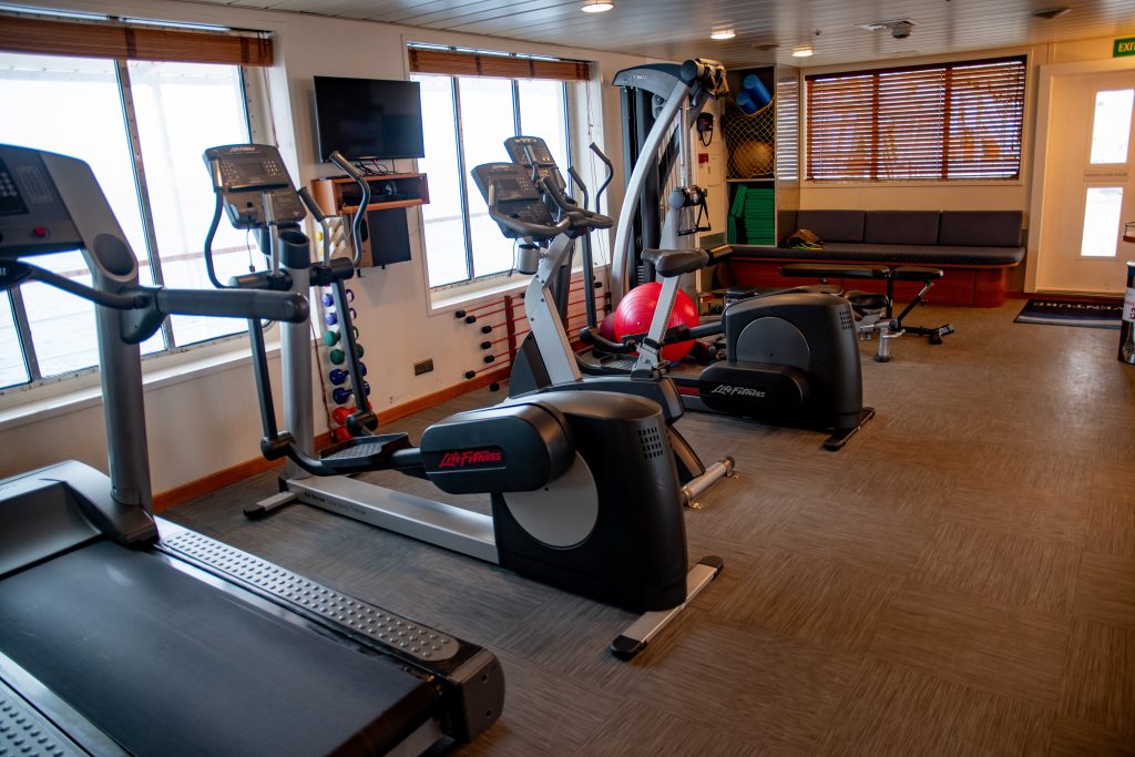 Fitness center on the ship Seaventure showing treadmill, ellipticals, weight machine, large windows and bench seating.  