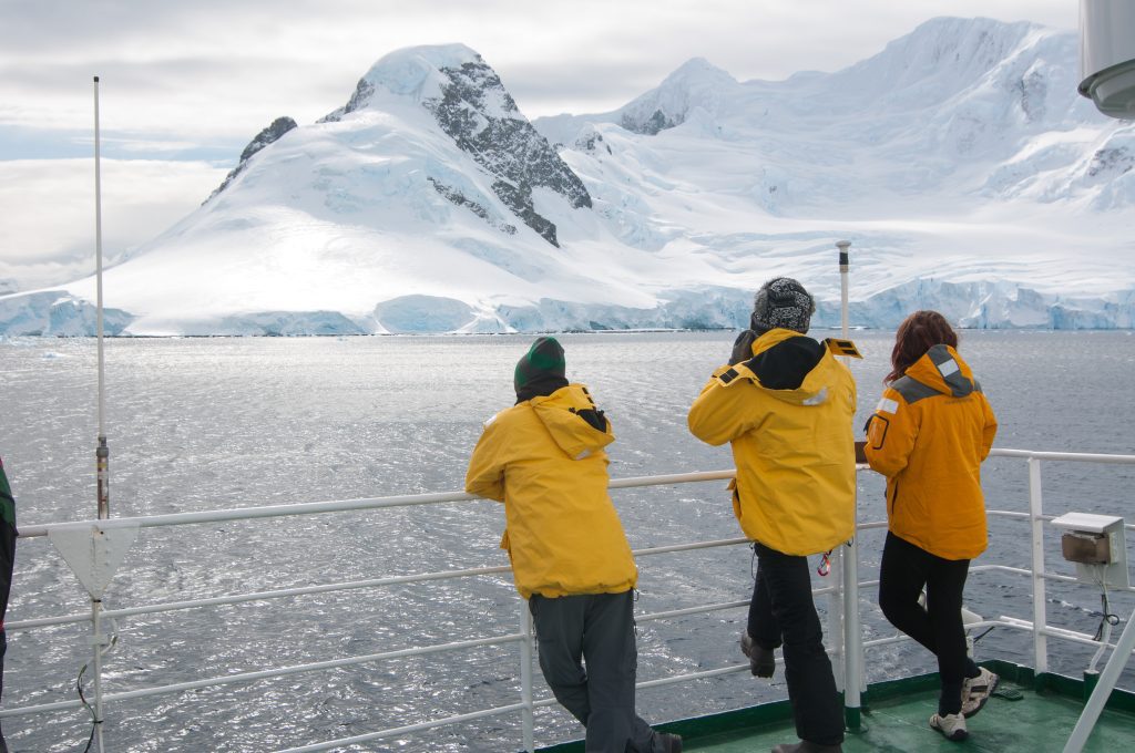 Three passengers standing on deck looking at snow covered mountain. 
