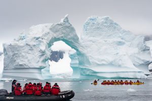 Zodiac and kayaks on the water in front of large arch iceberg. 