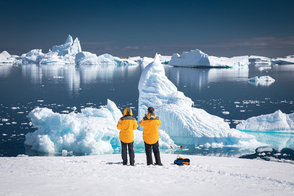 Two guest standing on snowy shoreline looking at icebergs. 