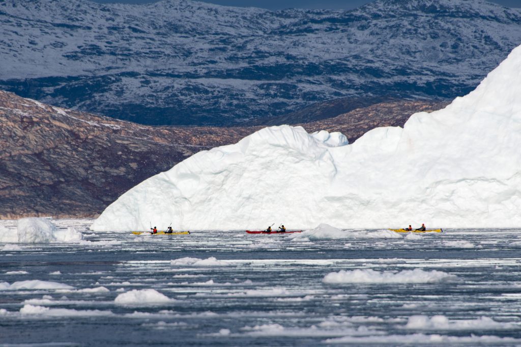 Kayaks on the water paddling next to large iceberg.