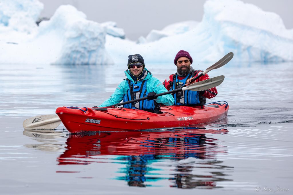 Two guests kayaking on a double kayak with a large iceberg in the background.