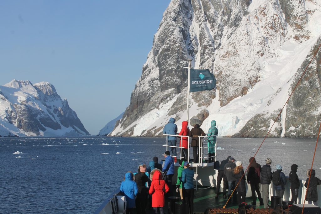 Passengers standing at bow of ship looking at rugged mountain. 
