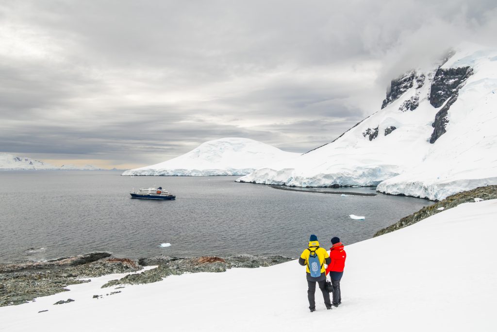 Two guests standing on snowy hillside looking at the ship Hondius. 