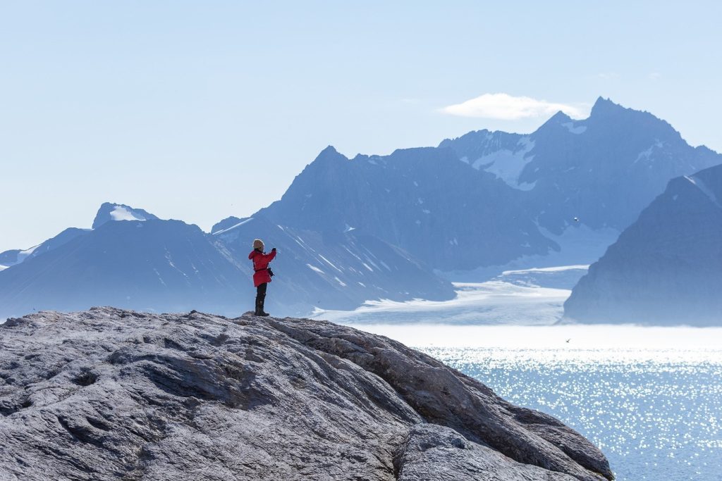 A guest standing at top of a rock ledge with ocean & mountains in background.