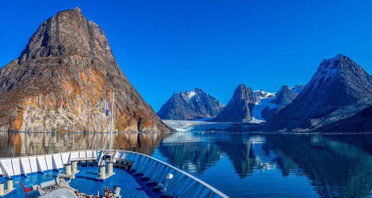 Ship bow sailing toward glacier with steep mountain peaks on the Heart of the Arctic itinerary