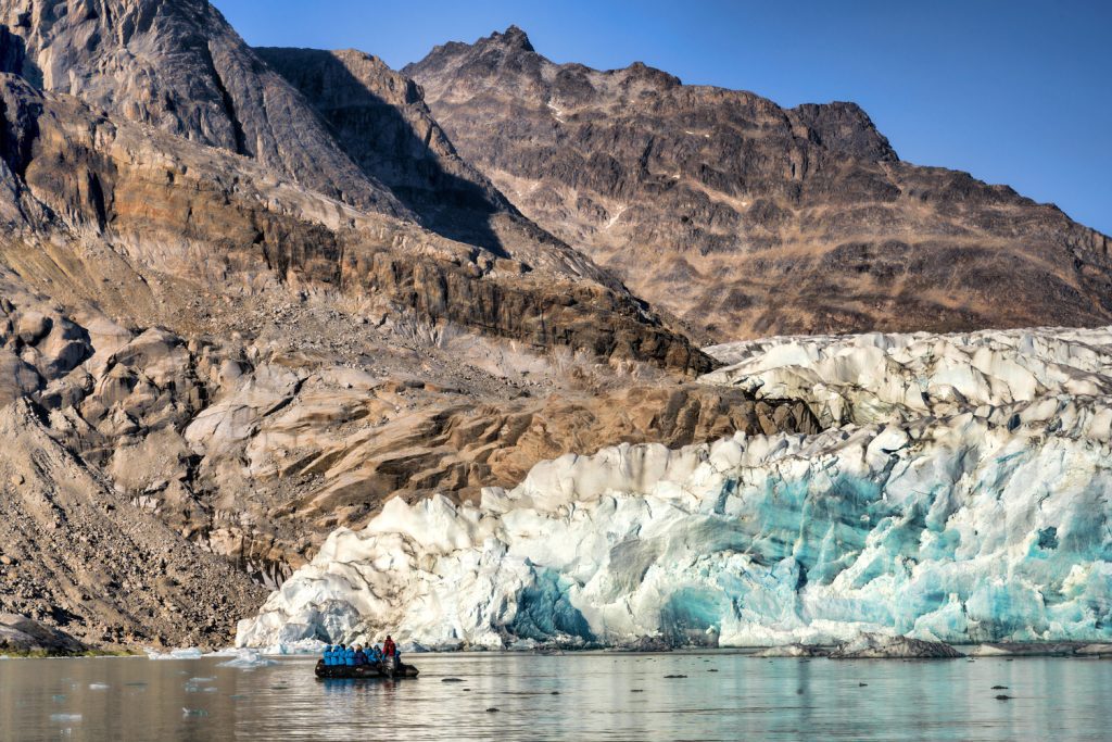 Zodiac on the water next to glacier face & rocky mountains.
