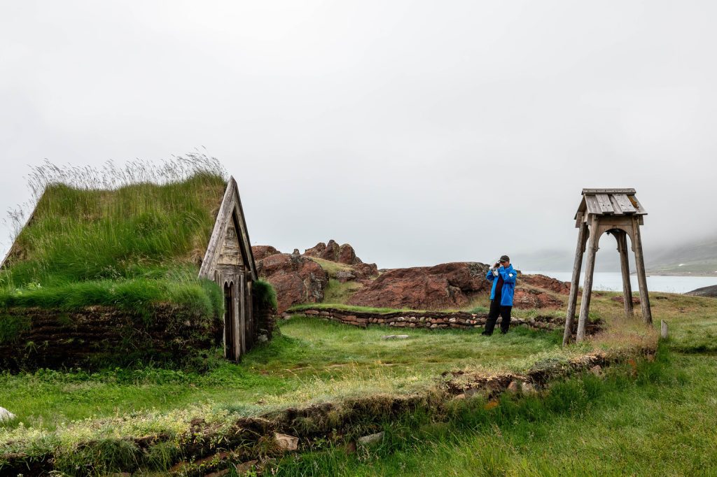 Guest standing next to old norse house with grass on roof. 