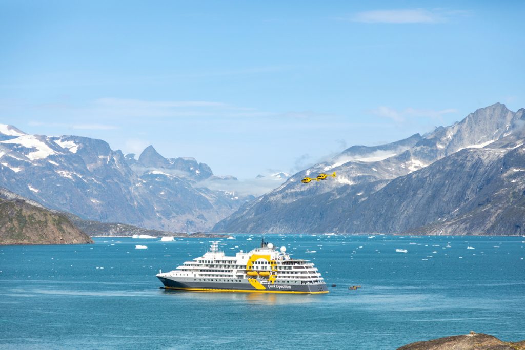 The ship Ultramarine in a scenic fjord with helicopters flying nearby.