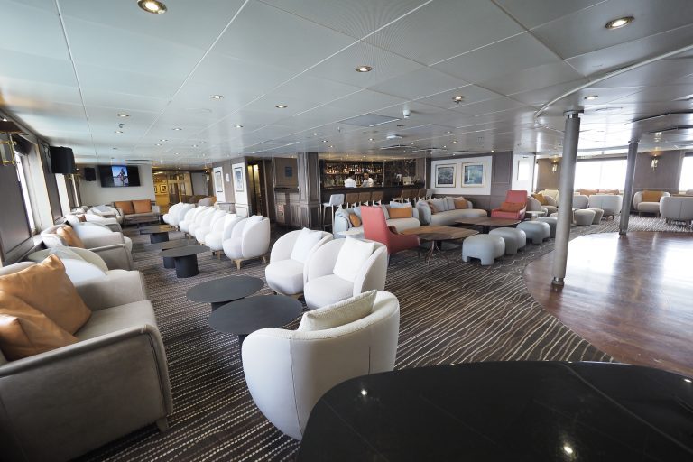 The main lounge onboard Exploris One with many chairs, coffee tables and a bar.