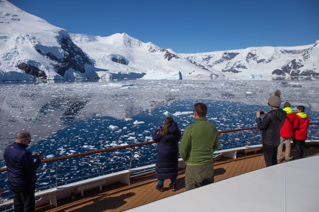 Passengers standing on World Voyager observation deck looking towards icy waters and snow covered mountains.