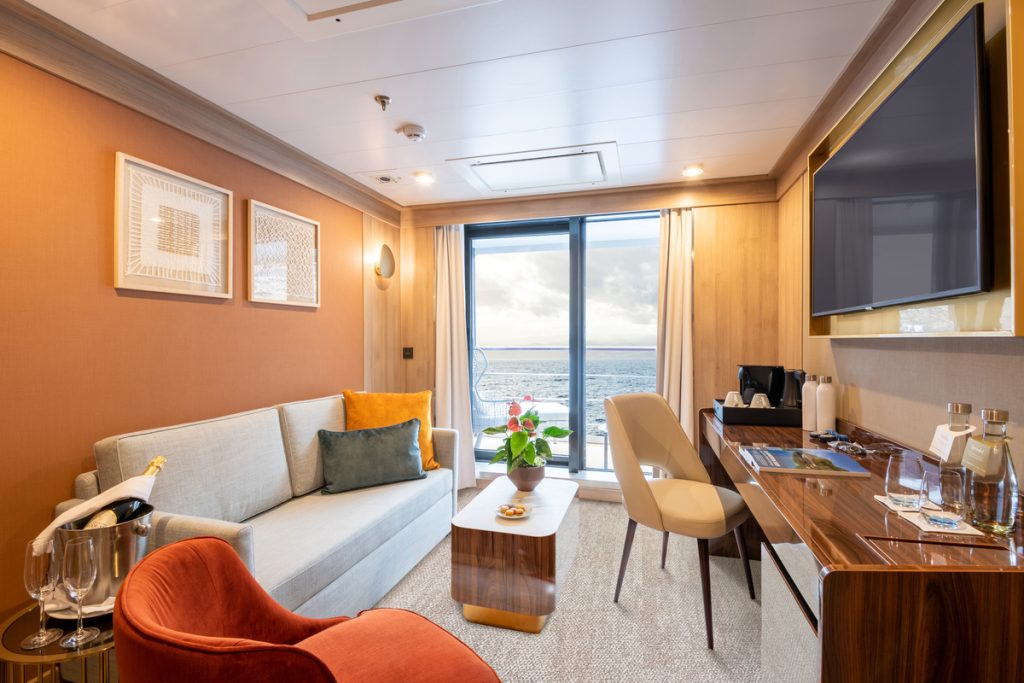 Sitting area in cabin with couch, desk, large window and balcony with sea view. 