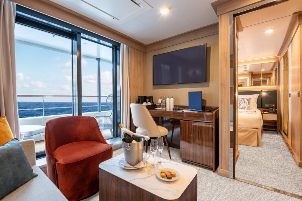 Owner's Suite on the ship World Voyager with desk, tv, door leading to separate bedroom and floor to ceiling glass windows. 