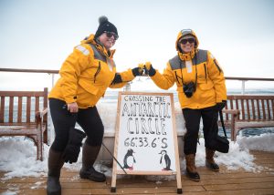 Two passengers standing next to Antarctic Circle sign on deck of a ship. 