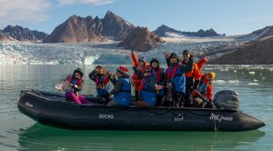 Guests in zodiac waving to camera with mountain & glacier in background.