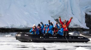 Guests on zodiac next to glacier waving at camera. 
