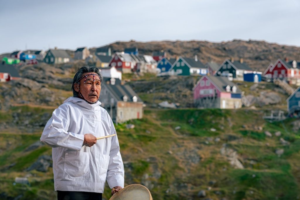 A local Greenlander with a drum standing in front of a village.