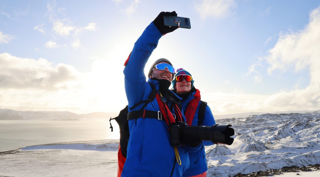 Two guests standing on shore in Antarctica taking a selfie. 
